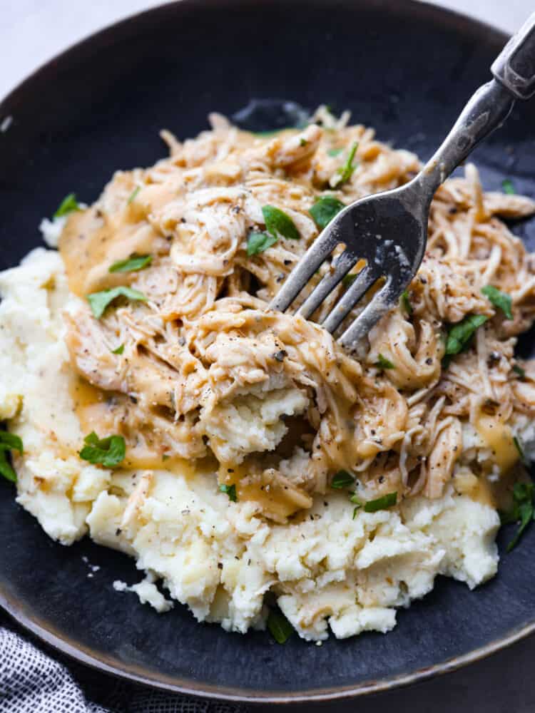 A close up shot of chicken and gravy on a pile of mashed potatoes on a black plate with a silver fork. 