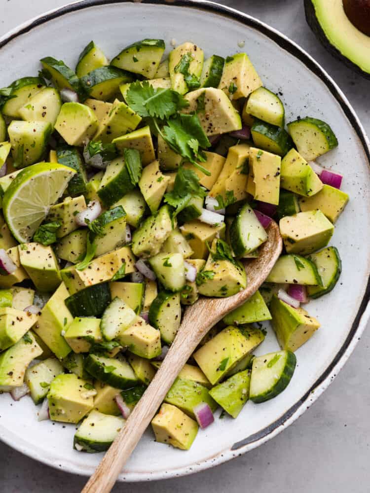 Cucumber and avocado salad in a white bowl with a wooden spoon.