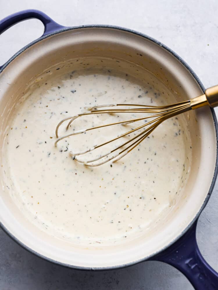 Close overhead photo of garlic parmesan sauce in a blue saucepan being stirred with a gold whisk.