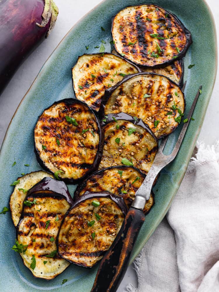 Top view of grilled eggplant on a blue oval shaped platter with a serving fork is on the platter.
