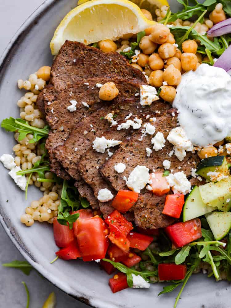 Closeup of a gyro bowl made with gyro meat slices, couscous, veggies, feta crumbles, and tzatziki sauce.