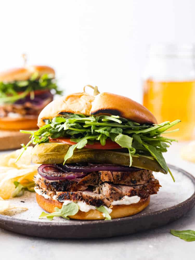 A pork tenderloin sandwich topped with pickles, tomatoes, greens, and grilled onions.