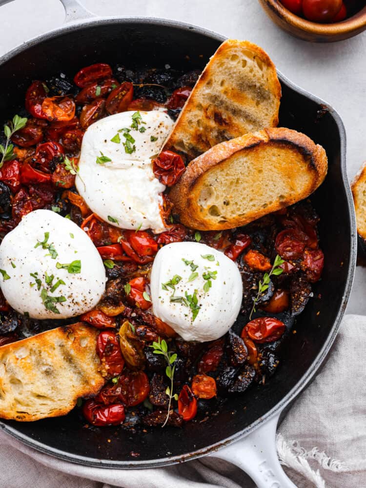 Top-down view of an titbit made with cherry tomatoes, figs, and burrata cheese. There are 2 pieces of toasted baguette in the skillet.