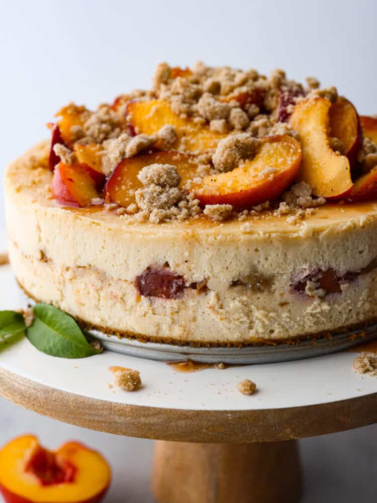 A peach cobbler cheesecake on a wooden and white cake stand. 