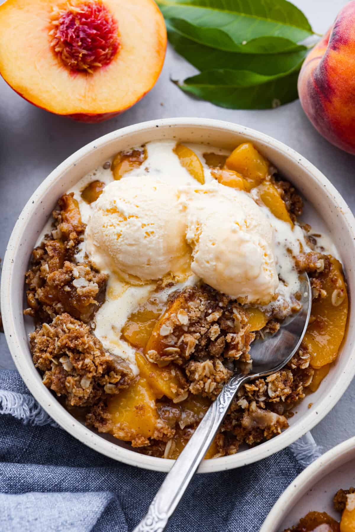 Peach crisp in a bowl with a silver spoon and a scoop of ice cream. 