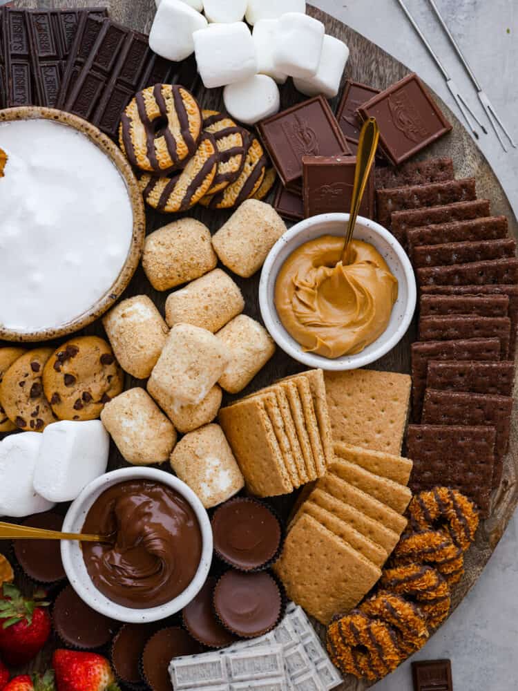 Closeup of a s’mores board. In addition to cookies, chocolate, and marshmallows, there are 2 small bowls filled with nutella and peanut butter.