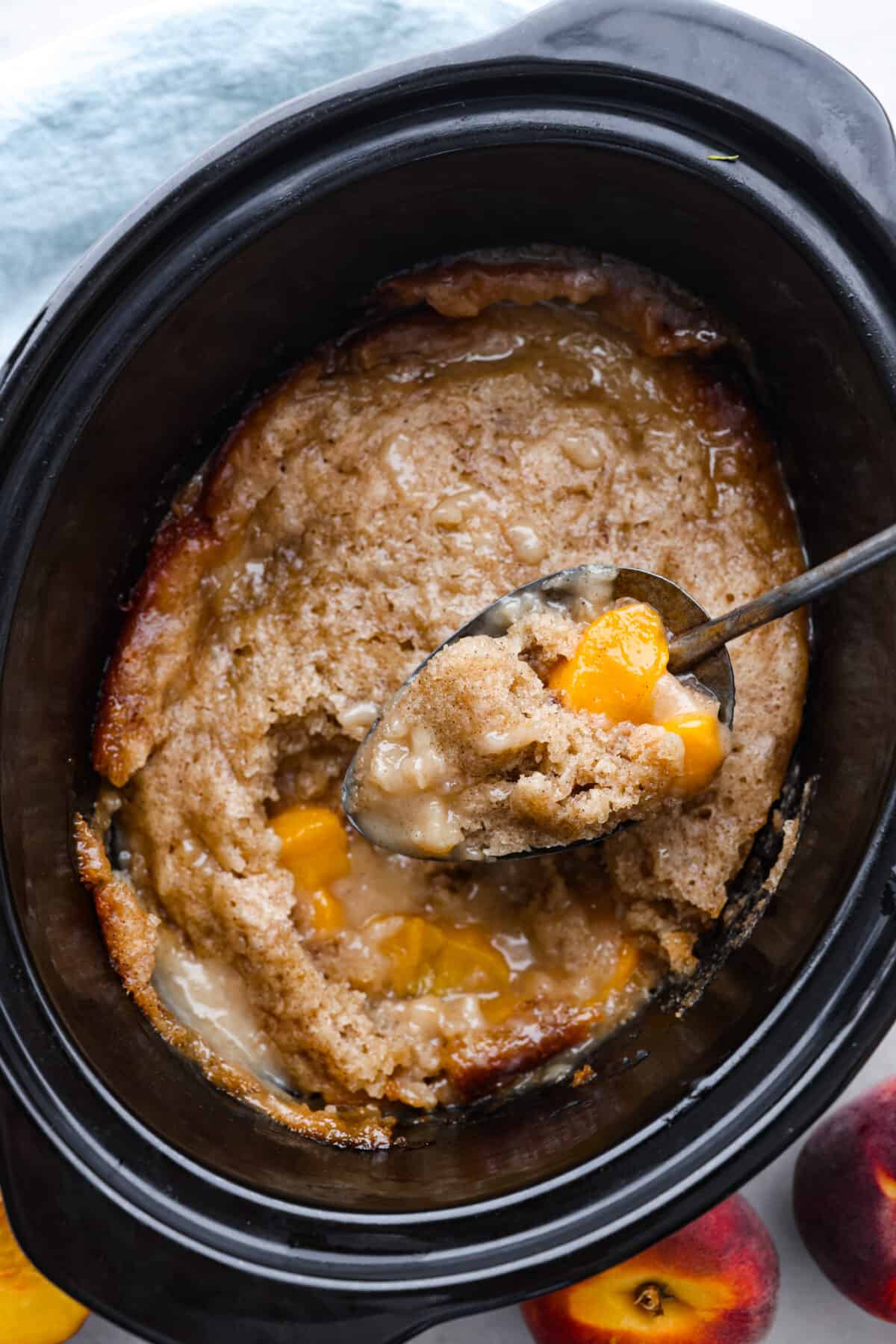 Peach cobbler in a slow cooker being scooped out with a spoon. 