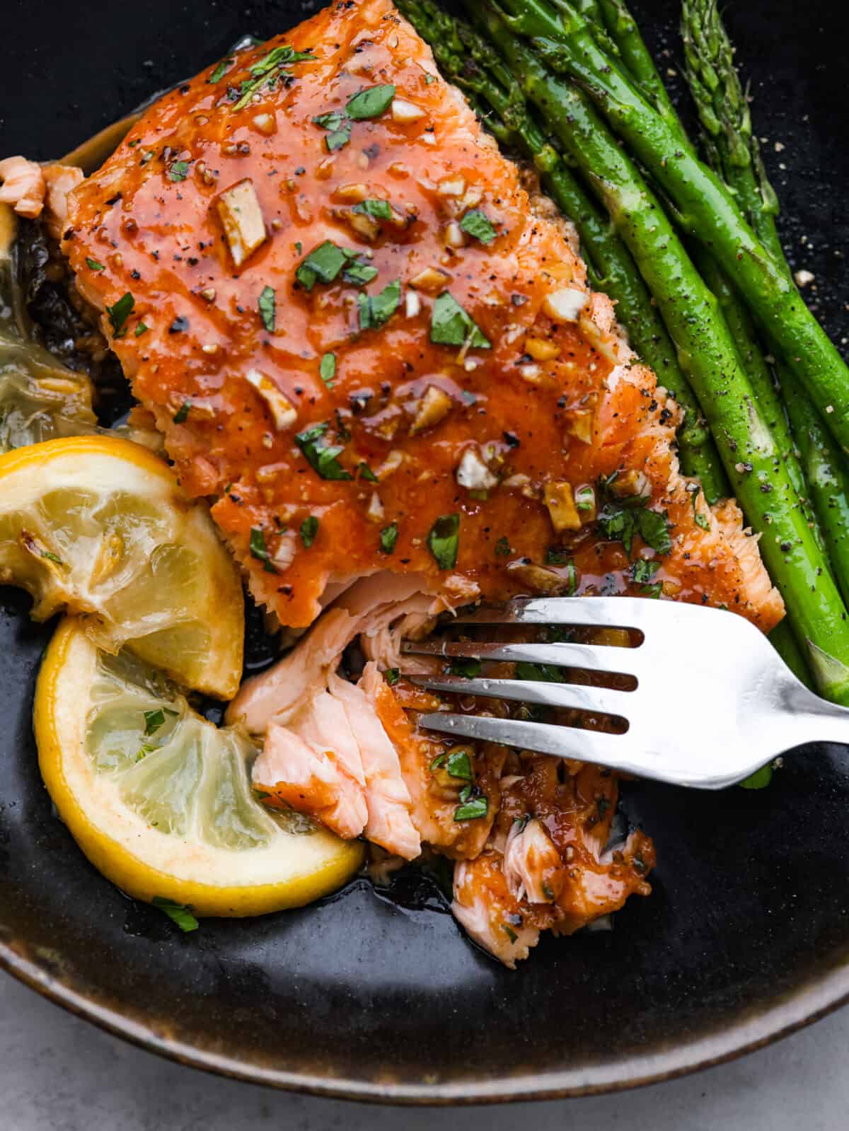 A close up of garlic salmon on a plate with asparagus and lemon slices being cut into with a fork. 