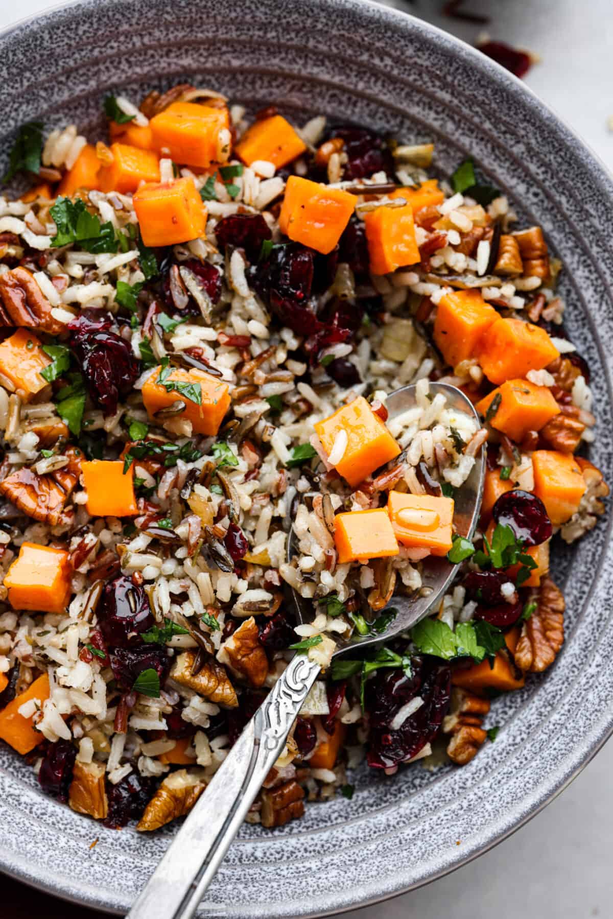 Close top view of cranberry pecan sweet potato wild rice pilaf in a gray bowl with a spoon. The spoon is lifting up the pilaf.