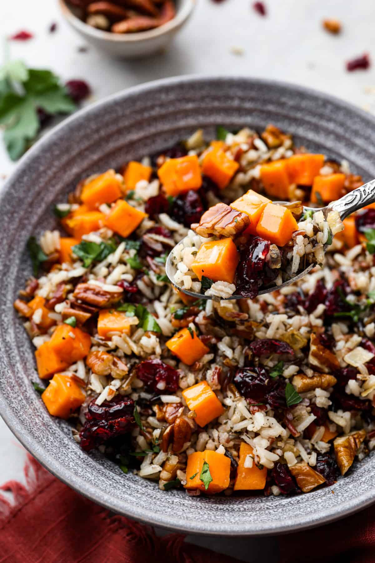 Side view of cranberry pecan sweet potato wild rice pilaf in a gray bowl with a spoon. The spoon is lifting up a bite of pilaf.