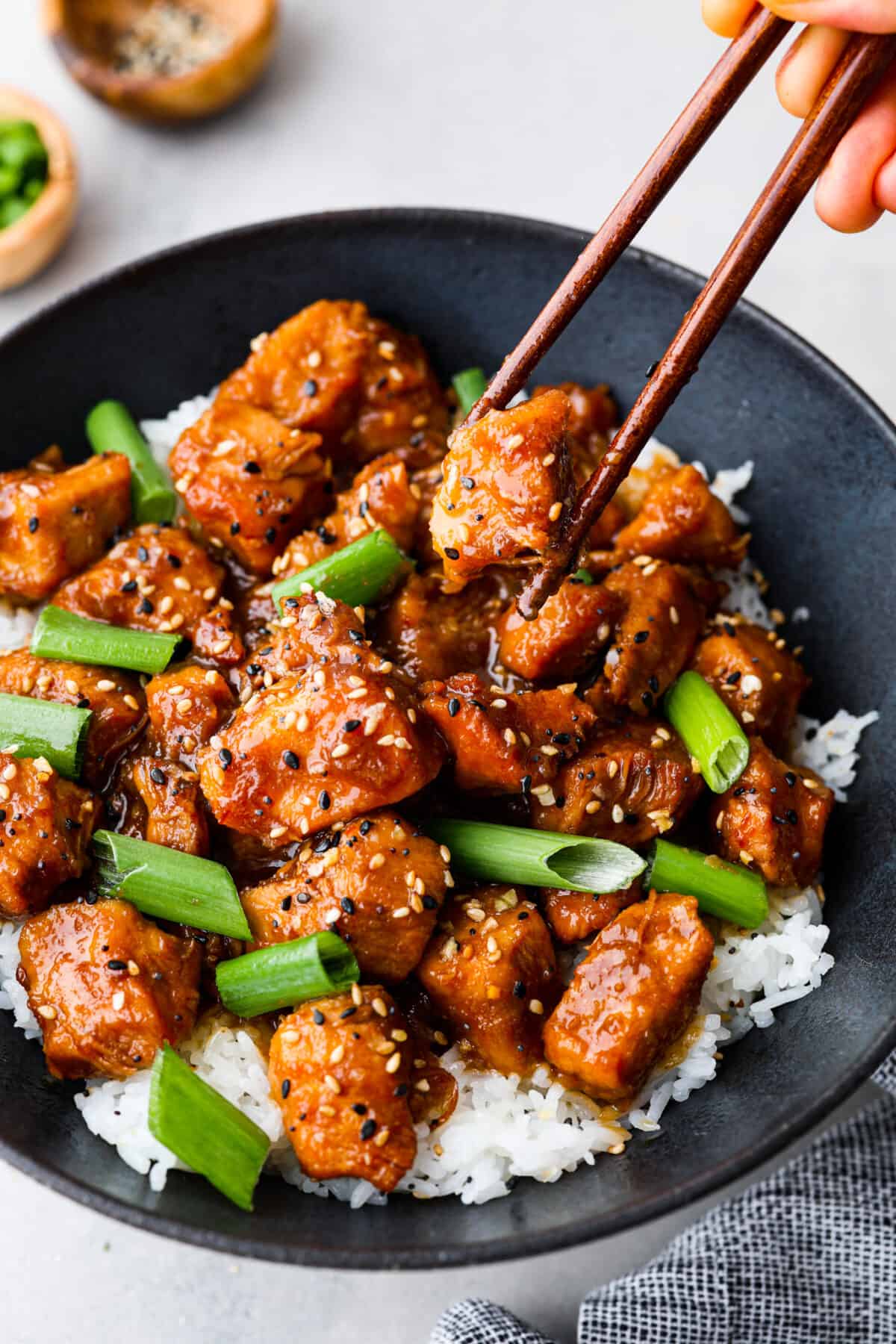 Close view of slow cooker General Tso's chicken in a black bowl served over rice. Garnished with sesame seeds and green onions. Chopsticks are picking up a piece of chicken.