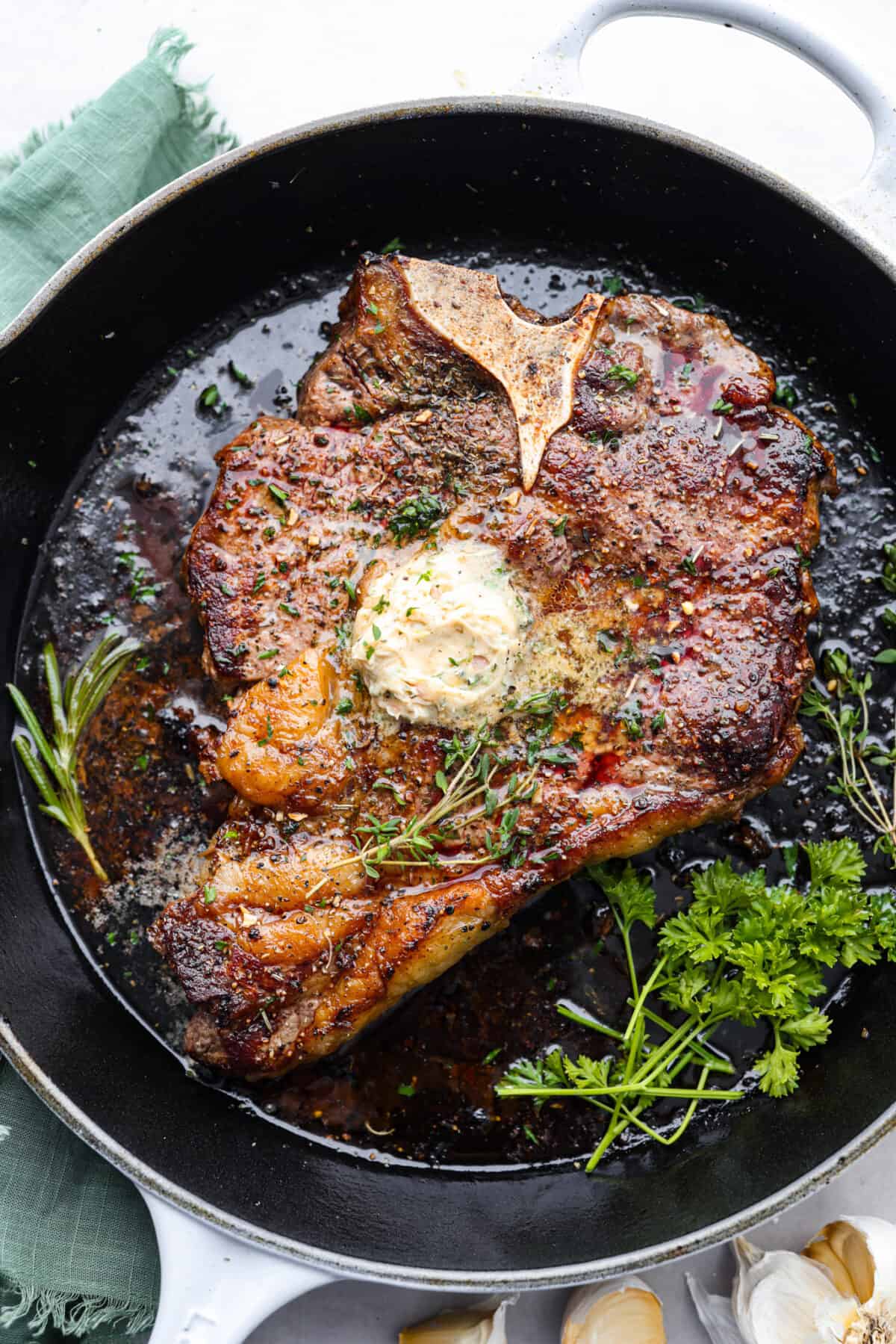 A cooked t-bone steak, topped with herbs and butter in a skillet.