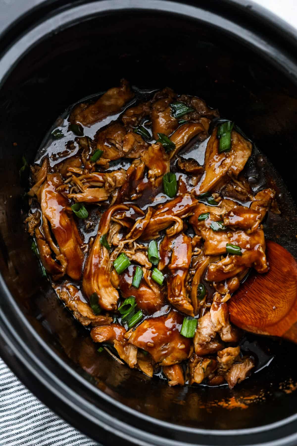 Top view of crockpot bourbon chicken in a black crockpot with a wooden spoon.
