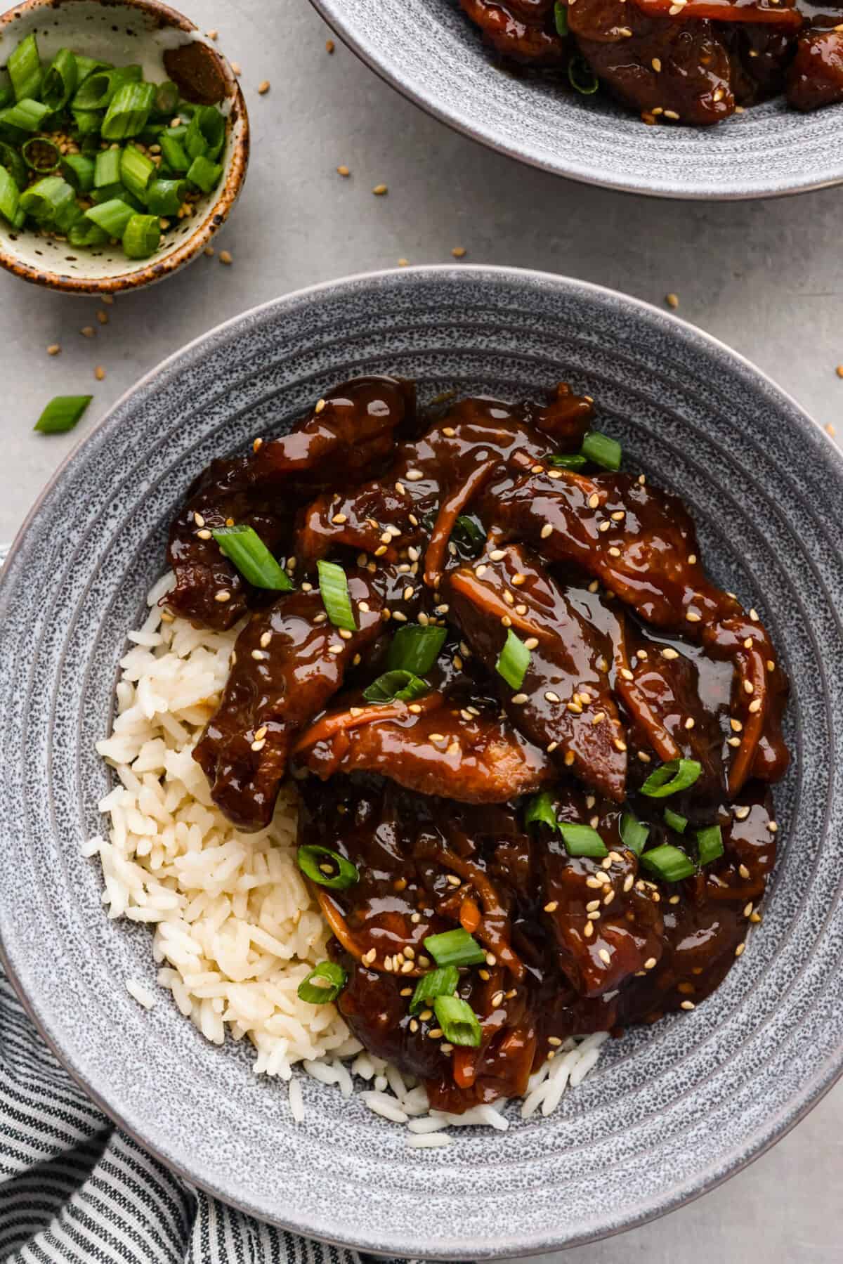 Overhead view of slow cooker Mongolian beef in a gray bowl over rice. Green onions and sesame seeds garnished on top.