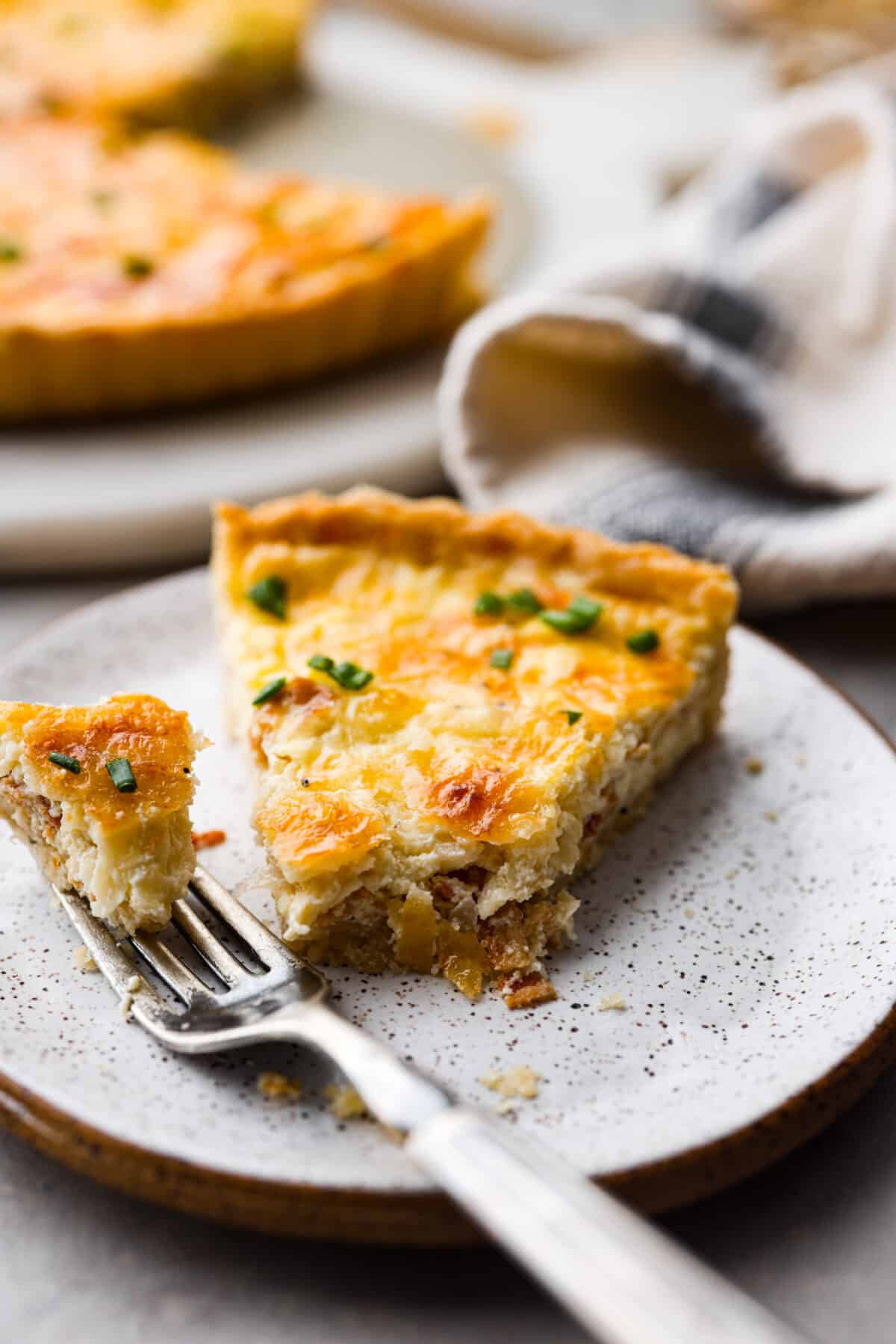 A slice of quiche lorraine with a bite taken out of it on a stoneware plate.