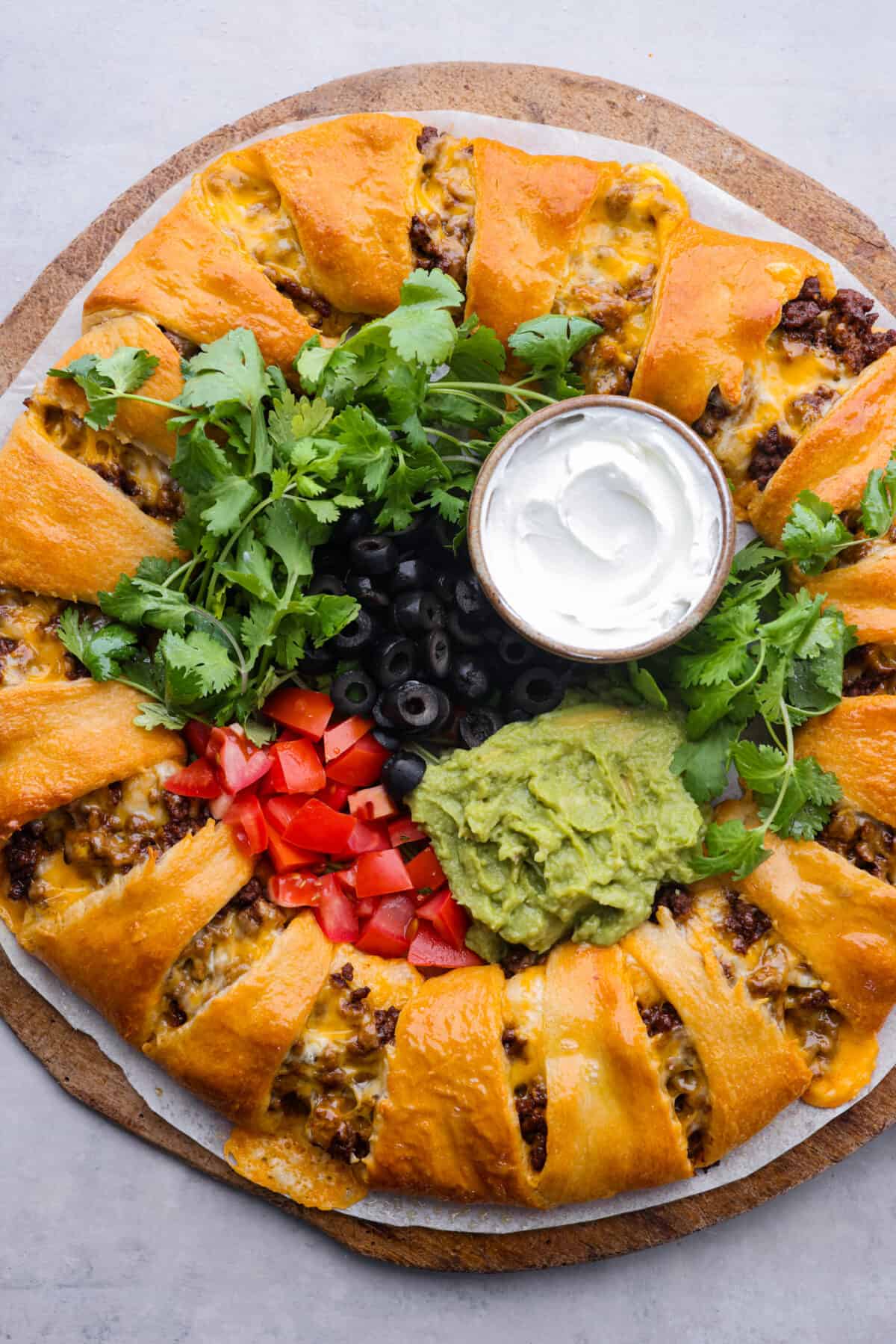 A whole taco ring, filled with servings of guacamole, sour cream, cilantro, tomatoes, and olives.