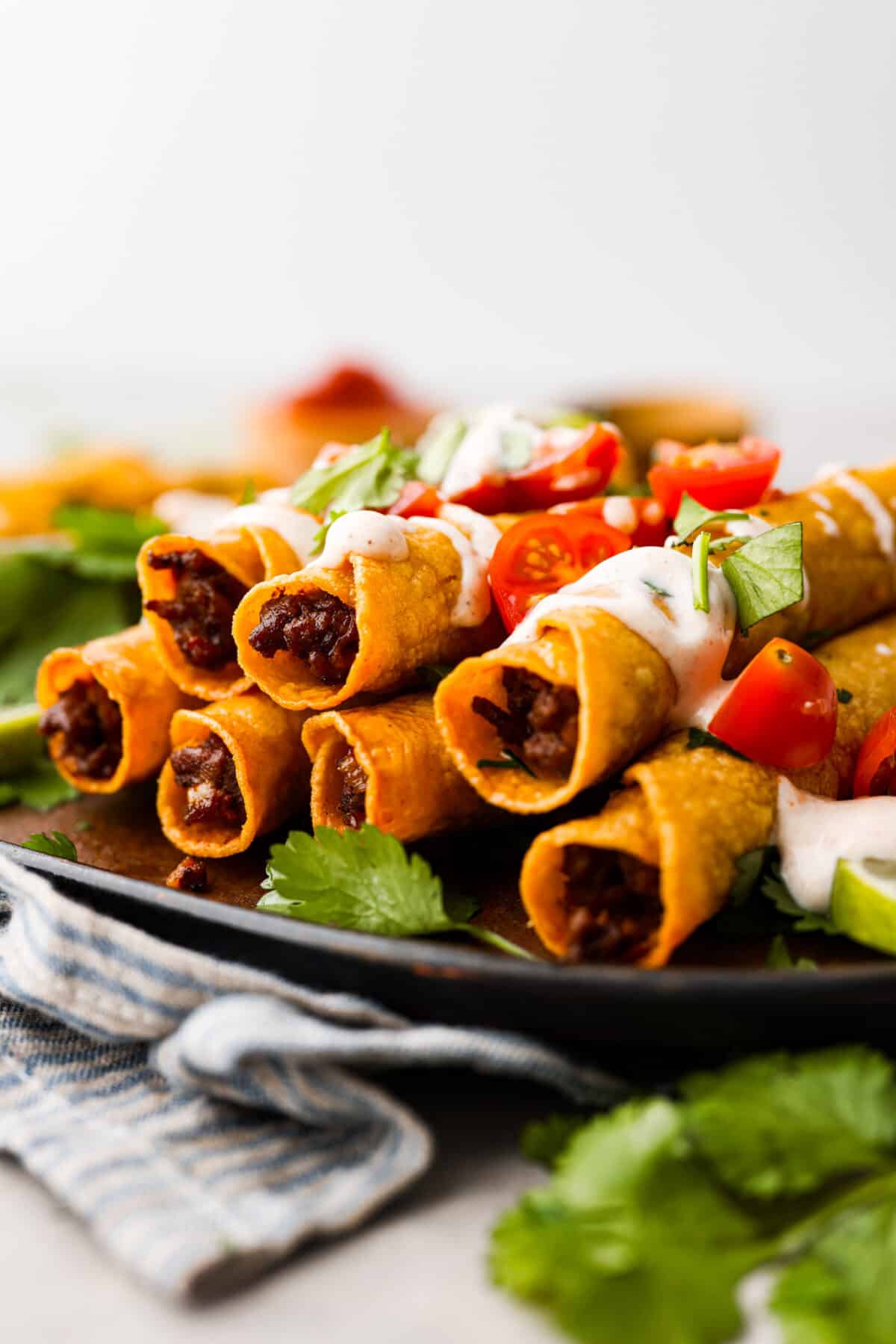 Beef taquitos on a black plate, topped with chopped tomatoes and sour cream.