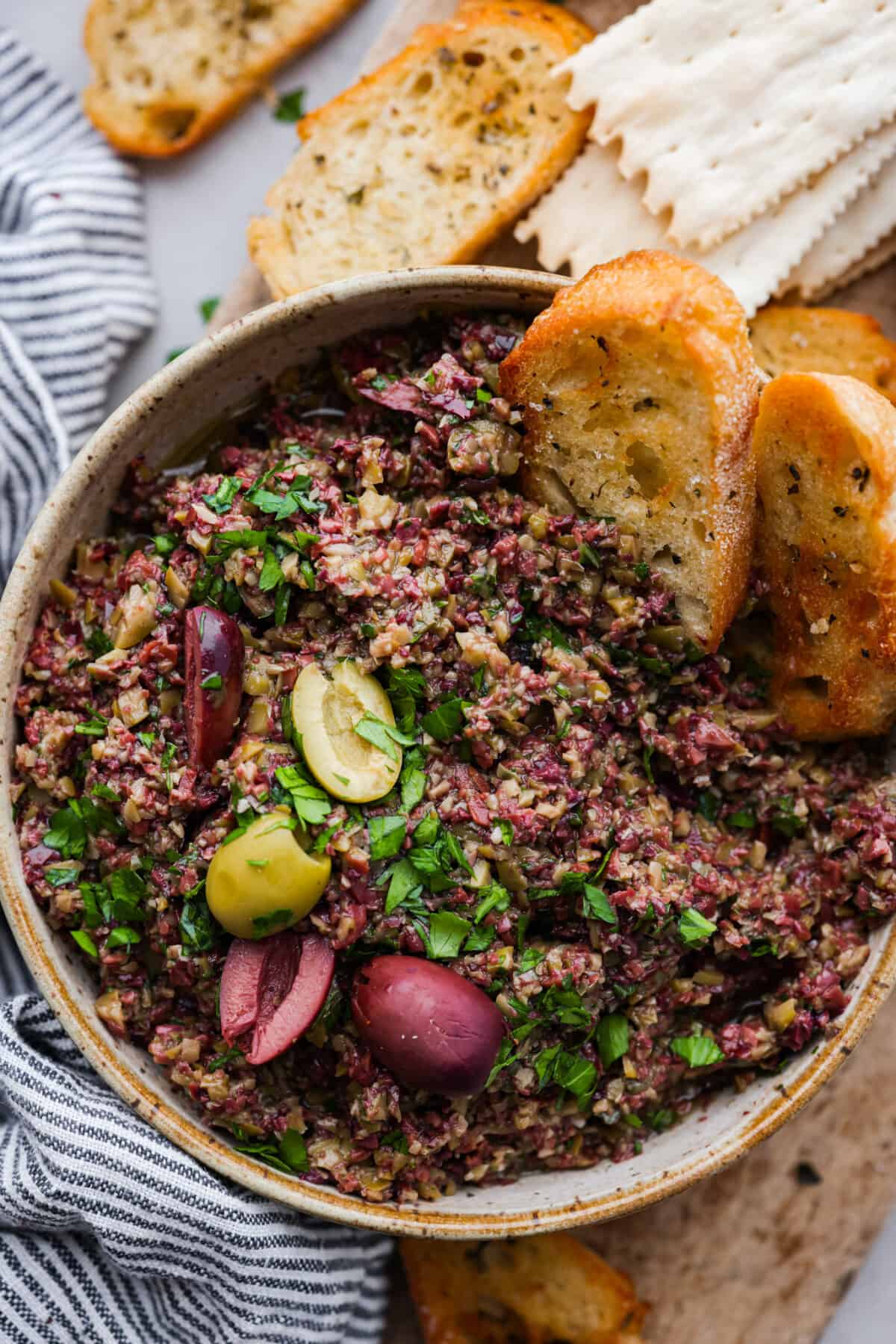 Close top view of olive tapenade in a bowl with crostini dipped in the spread. Olives and parsley are garnished on top.