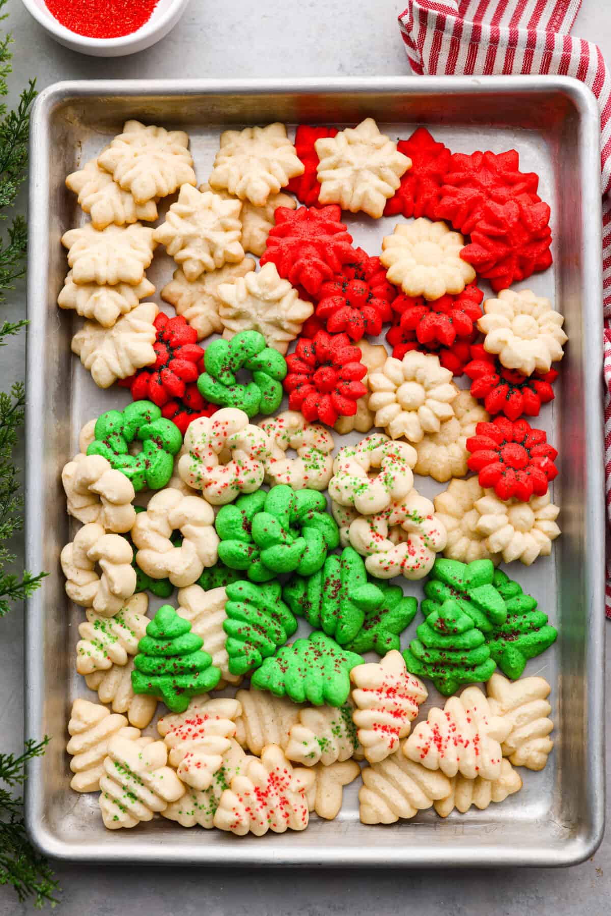 Spritz cookies in a baking sheet. Some are dyed green or red. They are in the shape of Christmas trees and snowflakes.