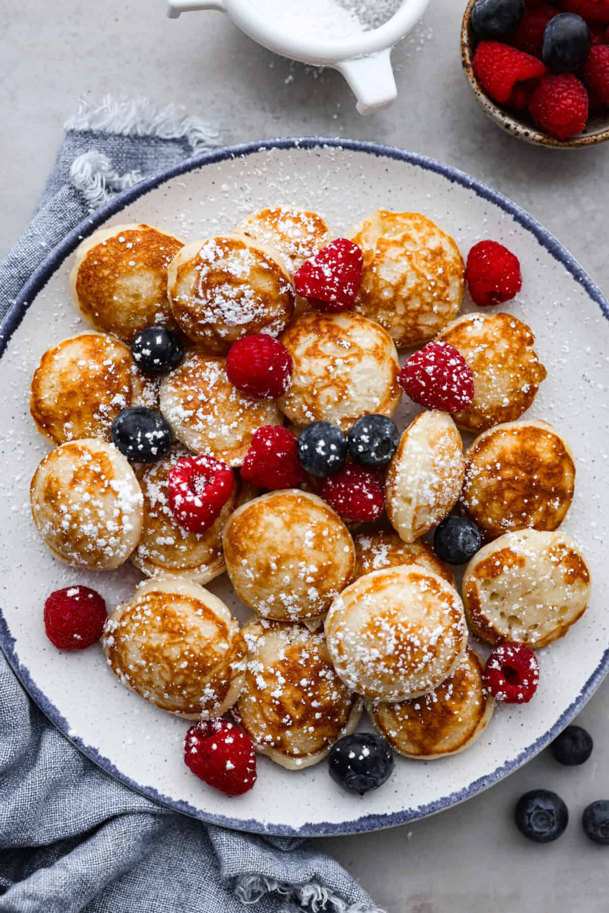 Poffertjes served with berries and a dusting of powdered sugar on a blue and white plate.