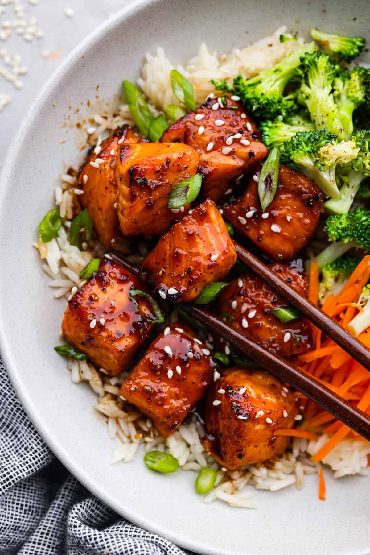 Say hello to your new favorite lunch: this honey-glazed salmon bowl made with juicy air fryer salmon bites, broccoli, and shredded carrots over white rice! Picking up a piece of salmon with chopsticks.