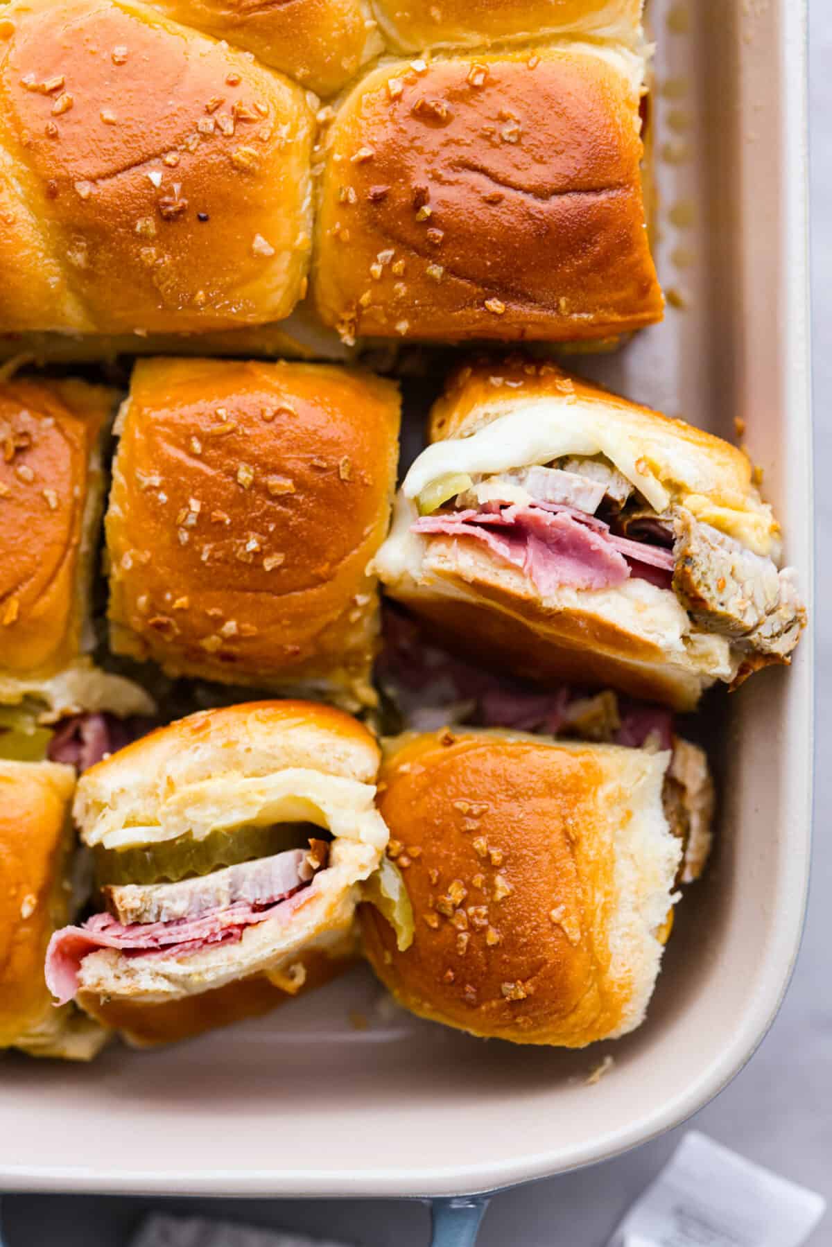 Top-down view of the Cuban sliders in a white baking dish. Two are turned on their side so you can see the filling.