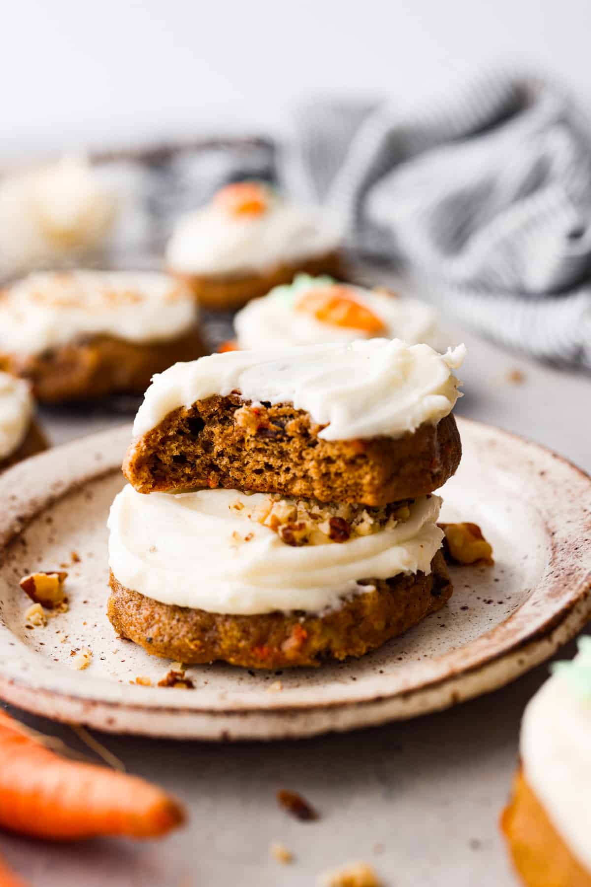 These frosted carrot cake cookies taste just like traditional carrot cake but in an easy-to-make cookie form! And it wouldn't be carrot cake without a generous amount of homemade cream cheese frosting on top. The perfect Spring dessert! 2 frosted carrot cake cookies stacked on top of each other. One has a bite taken out of it.