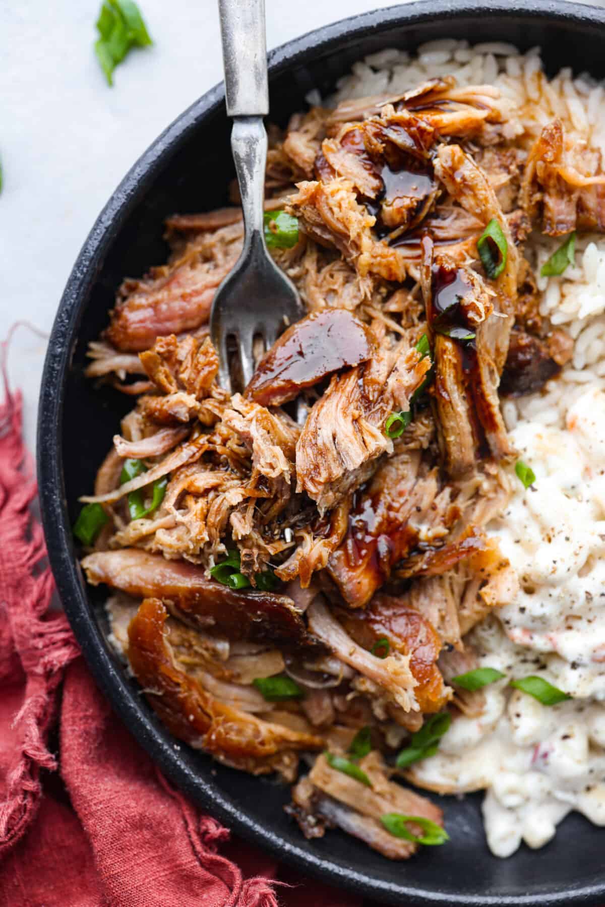 Top view of kalua pulled pork in a black bowl with rice and macaroni salad on the side. A fork is inside the bowl with teriyaki sauce and green onions garnished on top.