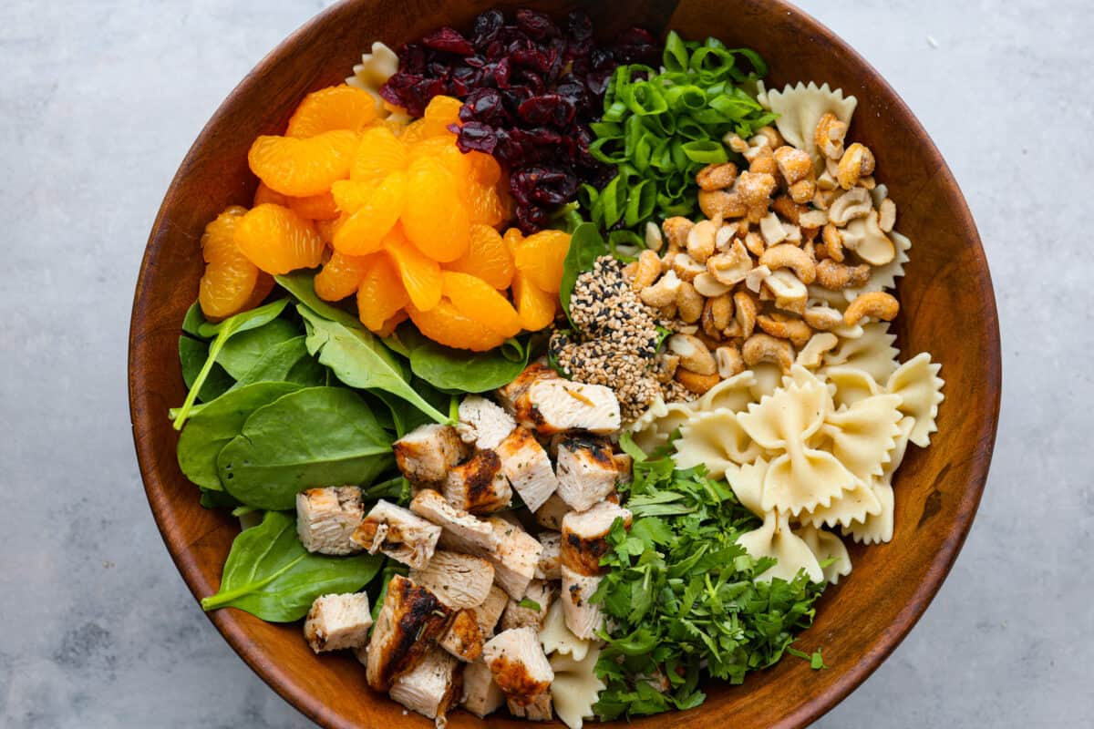 A wooden bowl filled with chicken, greens, mandarin oranges, bowtie pasta, and cashews.
