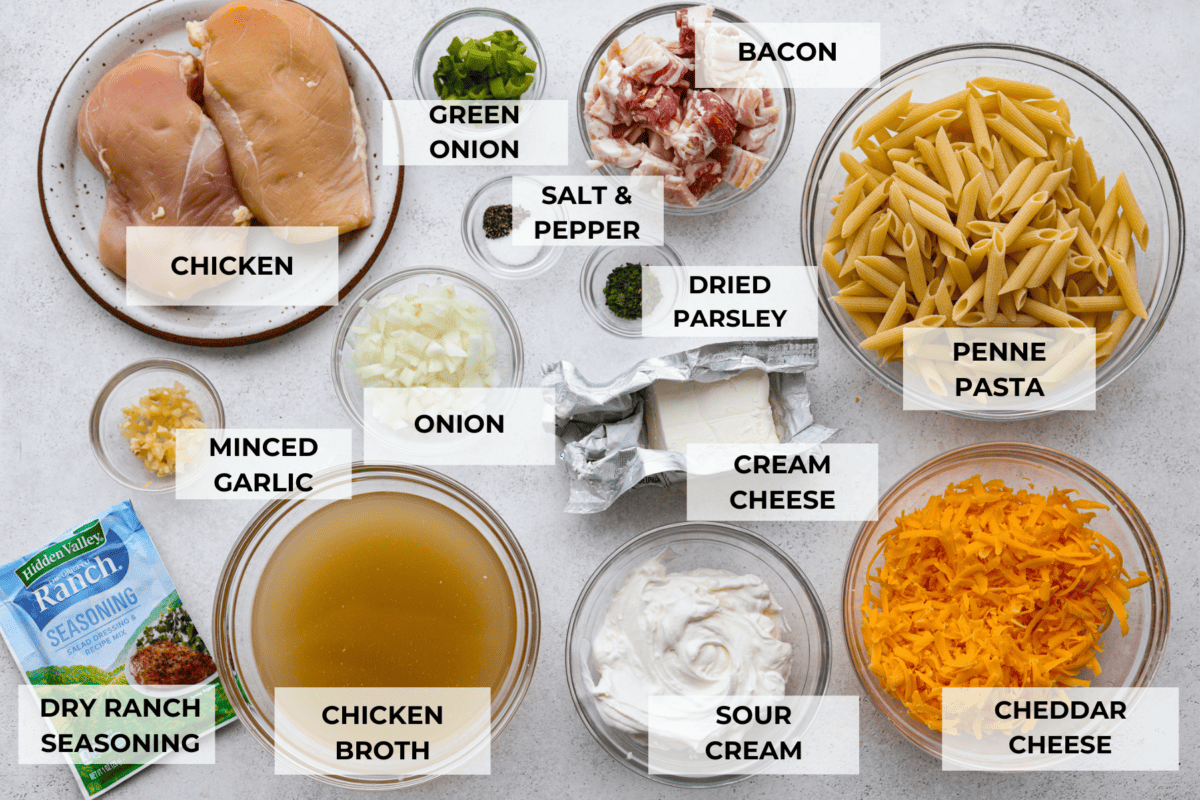All of the crack chicken pasta ingredients laid out in glass bowls. Listed are: chicken, green onion, bacon, salt and pepper, dried parsley, penne pasta, minced garlic, onion, cream cheese, dry ranch seasoning, chicken broth, sour cream, and cheddar cheese.