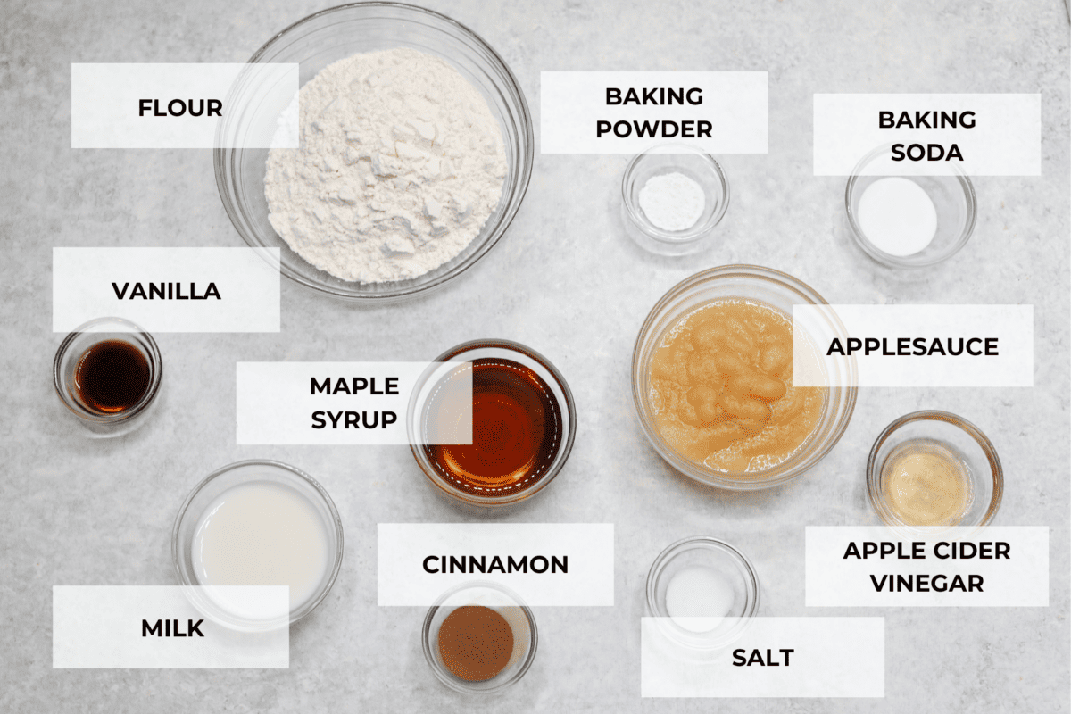 All of the muffin ingredients in glass bowls. Listed are: flour, baking powder, baking soda, vanilla, maple syrup, applesauce, milk, cinnamon, salt, and apple cider vinegar.