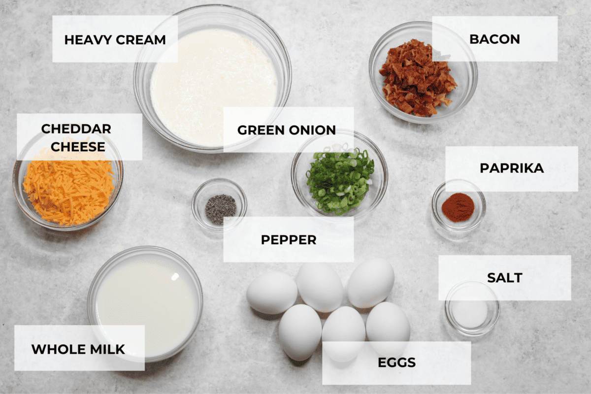 All of the ingredients laid out for crustless quiche. Listed are: cheddar cheese, heavy cream, green onion, bacon, paprika, pepper, salt, eggs, and whole milk.