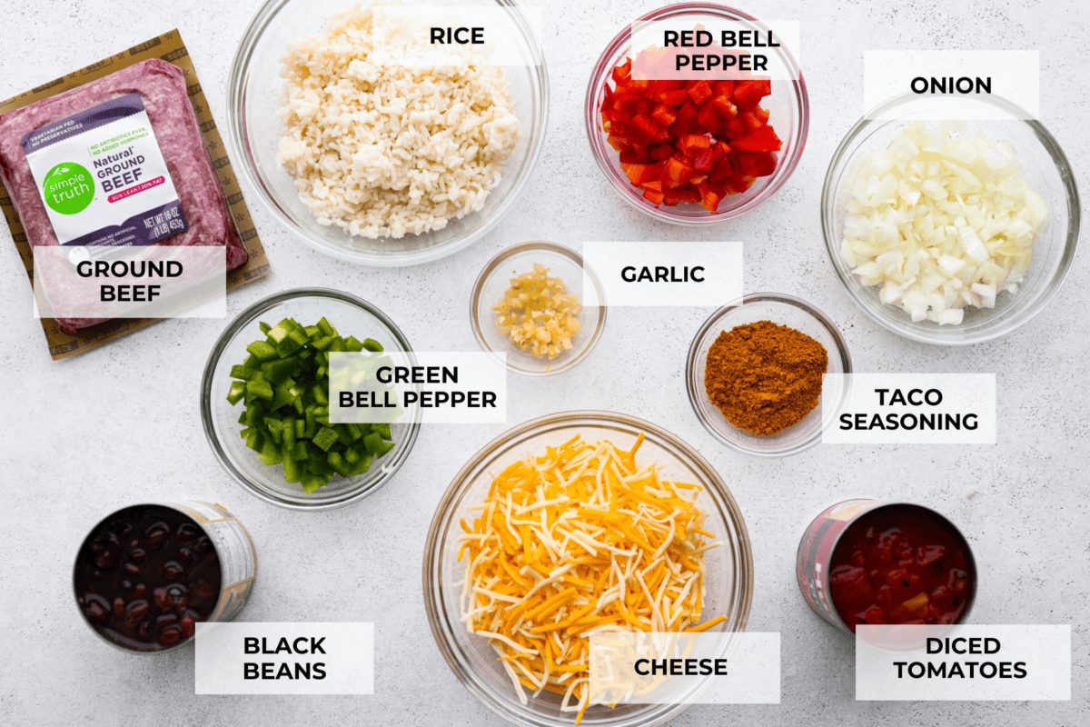 All of the ingredients for the taco skillet laid out in glass bowls. Listed are: ground beef, rice, green bell peppers, red bell peppers, garlic, taco seasoning, onion, black beans, cheese, and diced tomatoes.