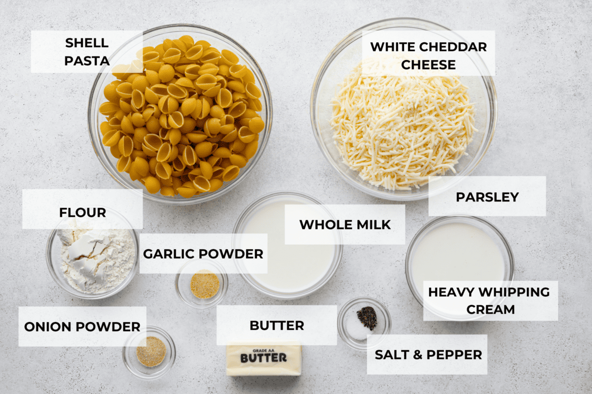 All of the ingredients for white cheddar mac and cheese in small glass bowls. Included are: shell pasta, white cheddar cheese, flour, garlic powder, whole milk, parsley, onion powder, butter, and salt and pepper.