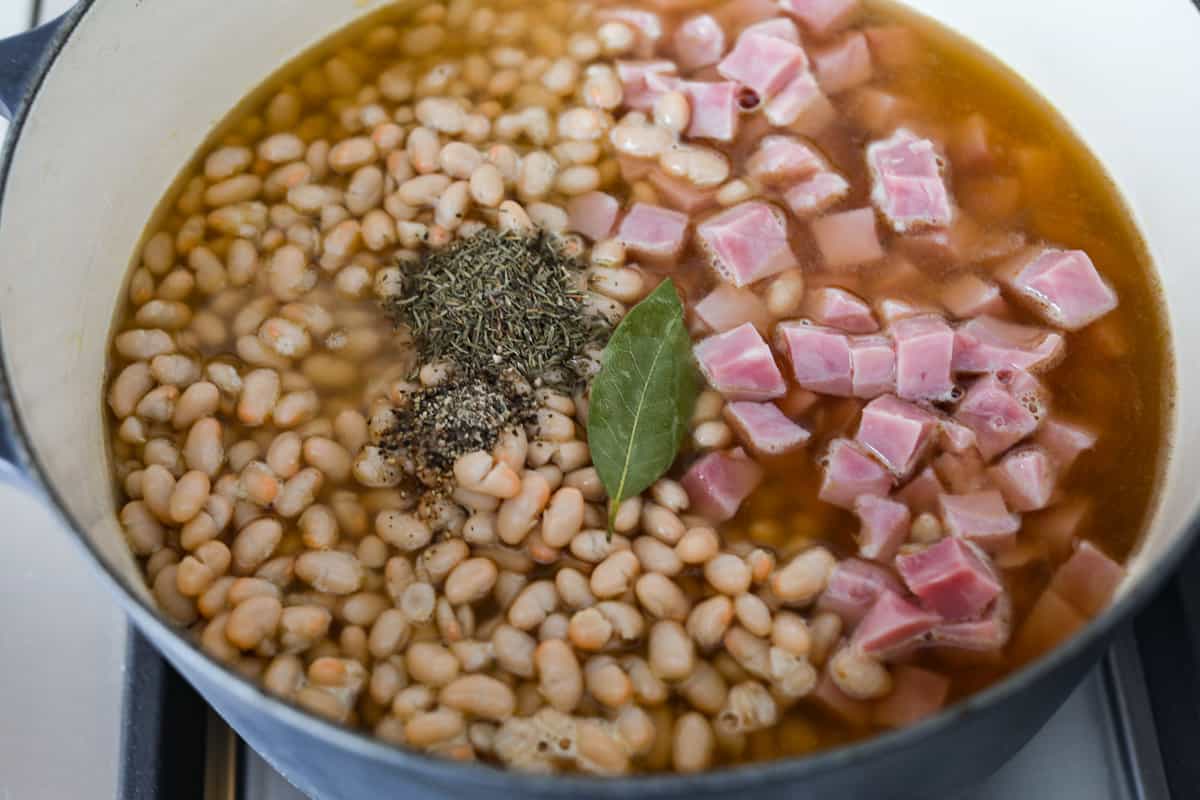 Comforting ham and bean soup is a classic and hearty soup. It's loaded with creamy white beans, smoky ham, and tender veggies. It's the perfect soup for using up your leftover ham and super easy to make on those busy weeknights. Fourth process photo of the bay leaf, pepper, and thyme added to the pot.