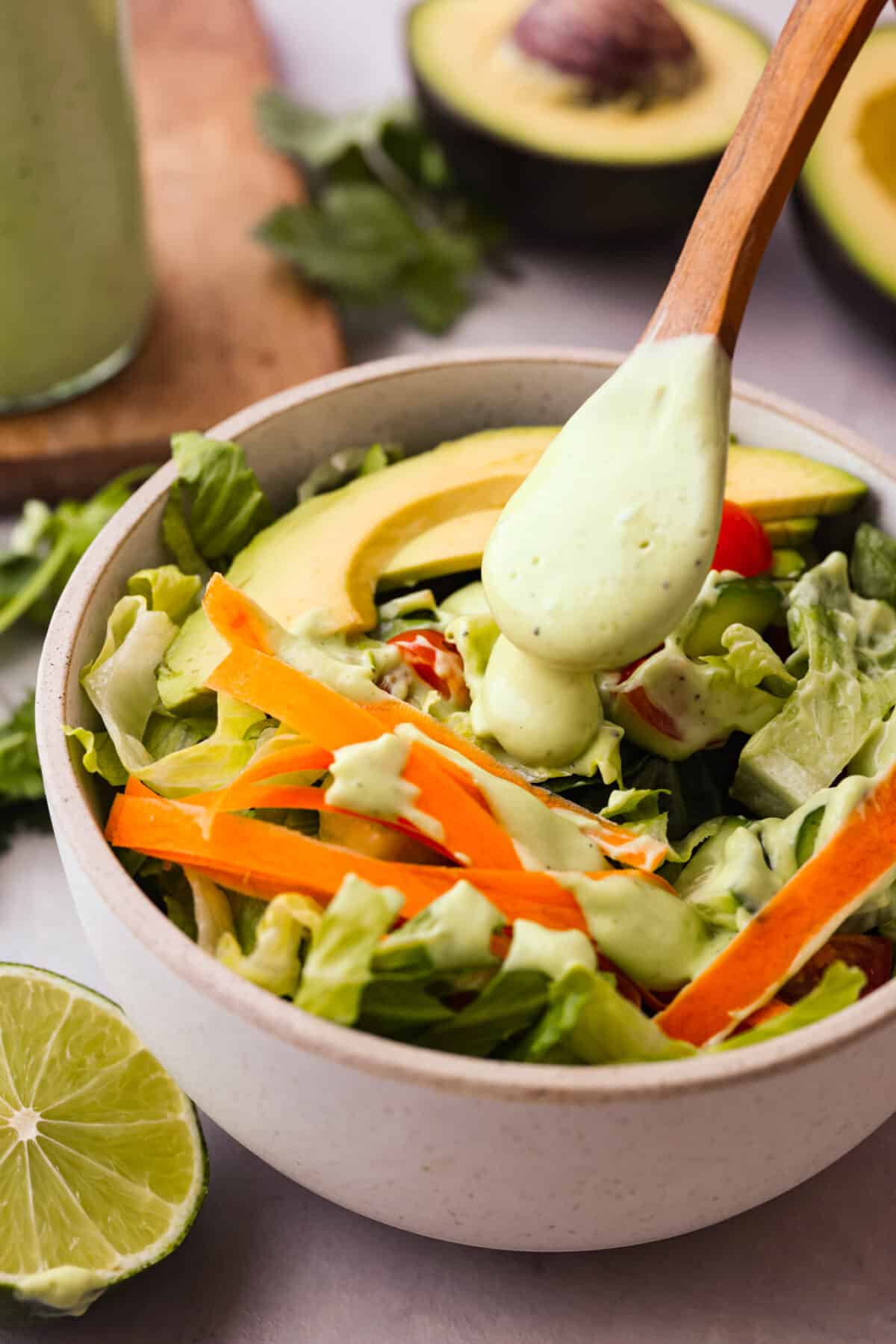 Side shot of a bowl of salad with veggies, avocado slices with a wooden spoon drizzling dressing over the top. 