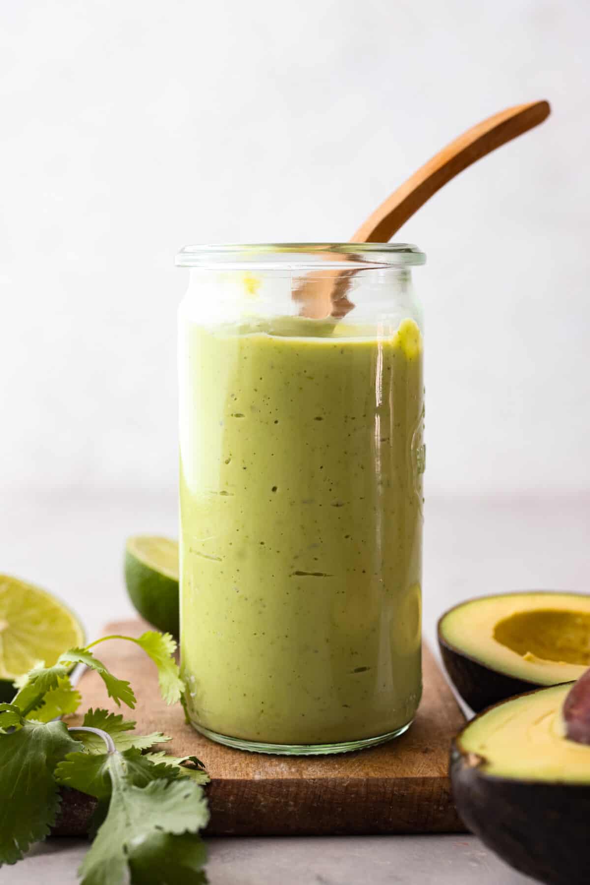 Side view of a glass jar filled with avocado dressing and a wooden spoon sticking out. There are avocados, limes and cilantro on the table next to the jar. 