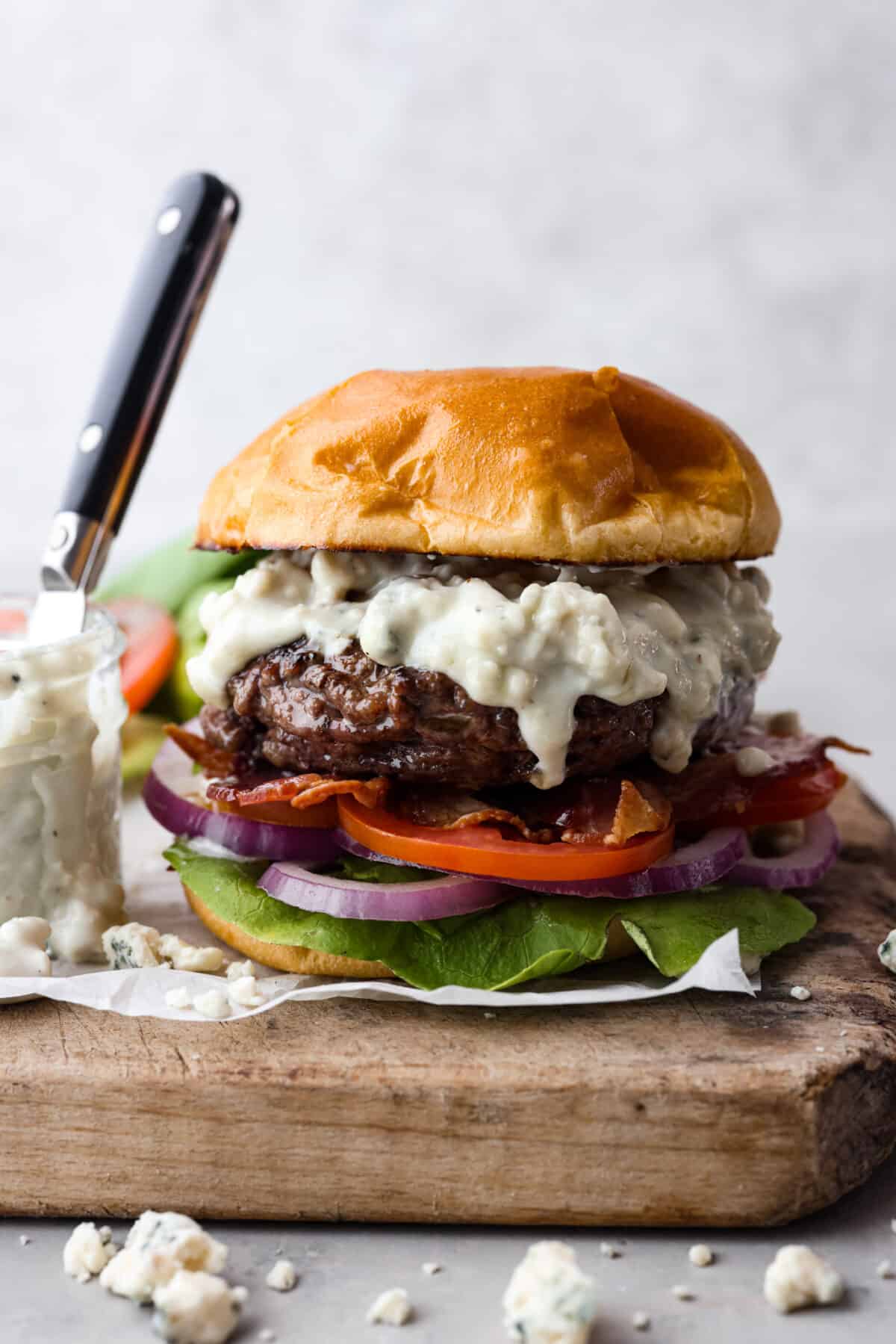 Side view of a burger with blue cheese dressing, bacon, lettuce tomato and onion on a crispy bun. All atop a wood cutting board with blue cheese crumbles. 