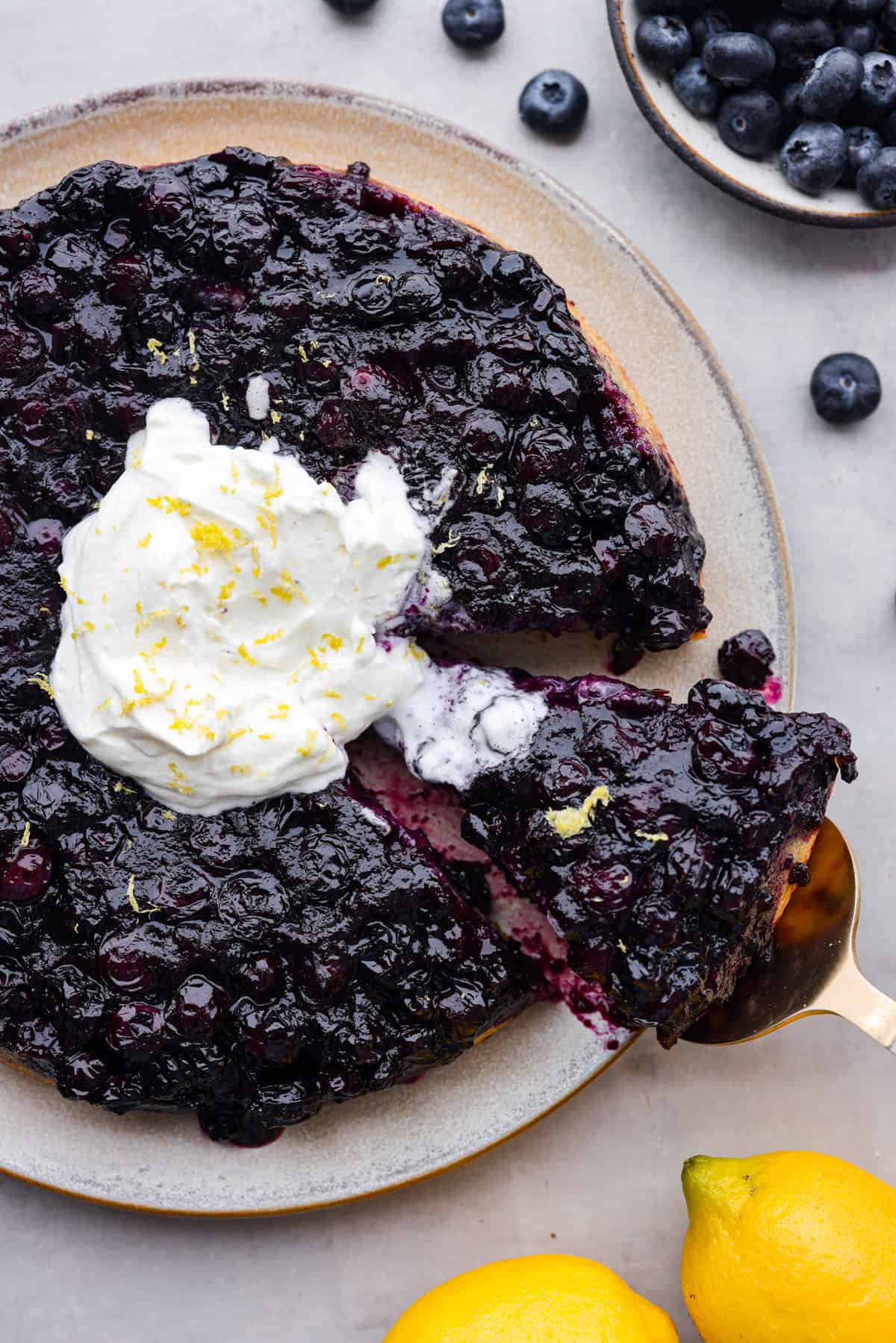 Be prepared to fall in love with this blueberry lemon upside-down cake! A delicious and moist lemon sponge cake bursting with juicy caramelized blueberries on top! Top view of blueberry lemon upside-down cake on a platter with whipped cream.