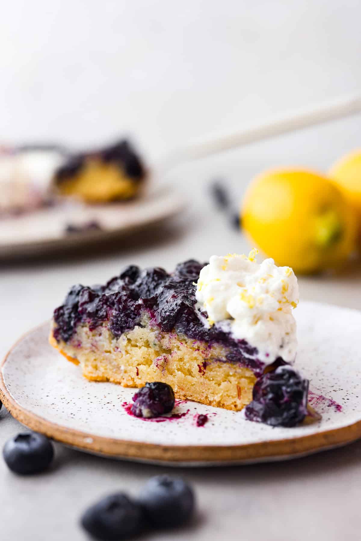 Be prepared to fall in love with this blueberry lemon upside-down cake! A delicious and moist lemon sponge cake bursting with juicy caramelized blueberries on top! Close view of a slice of blueberry lemon upside-down cake on a plate.