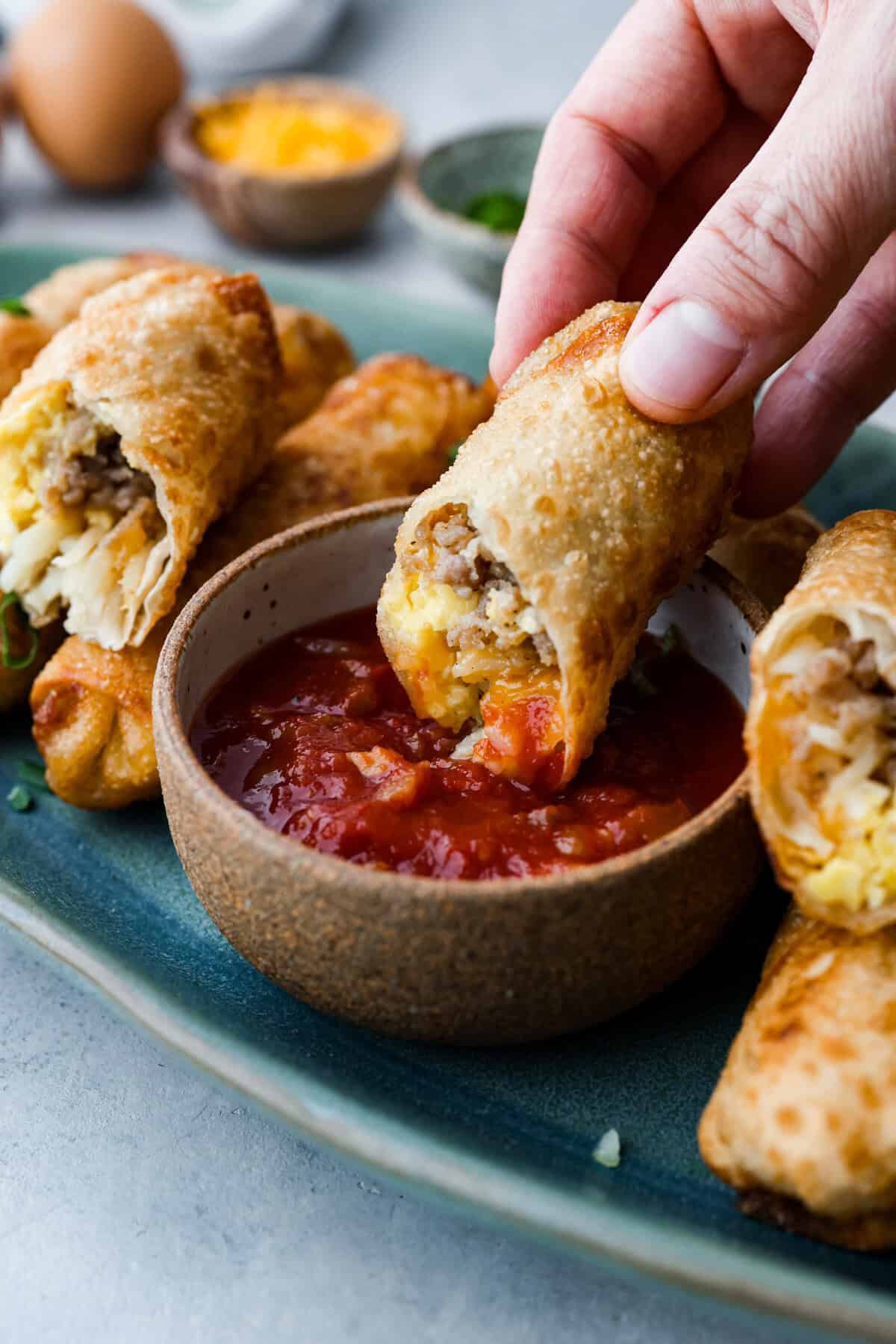 Breakfast Egg Rolls are a fun and delicious way to change up breakfast! The inside is filled with savory sausage, creamy eggs, hash browns, and plenty of cheese. Fry them up for a gooey, tender, savory center and a delicately crunchy exterior. This is a fun and unique breakfast that everyone will love! Side view of someone holding half of a breakfast egg roll dipping it into salsa.