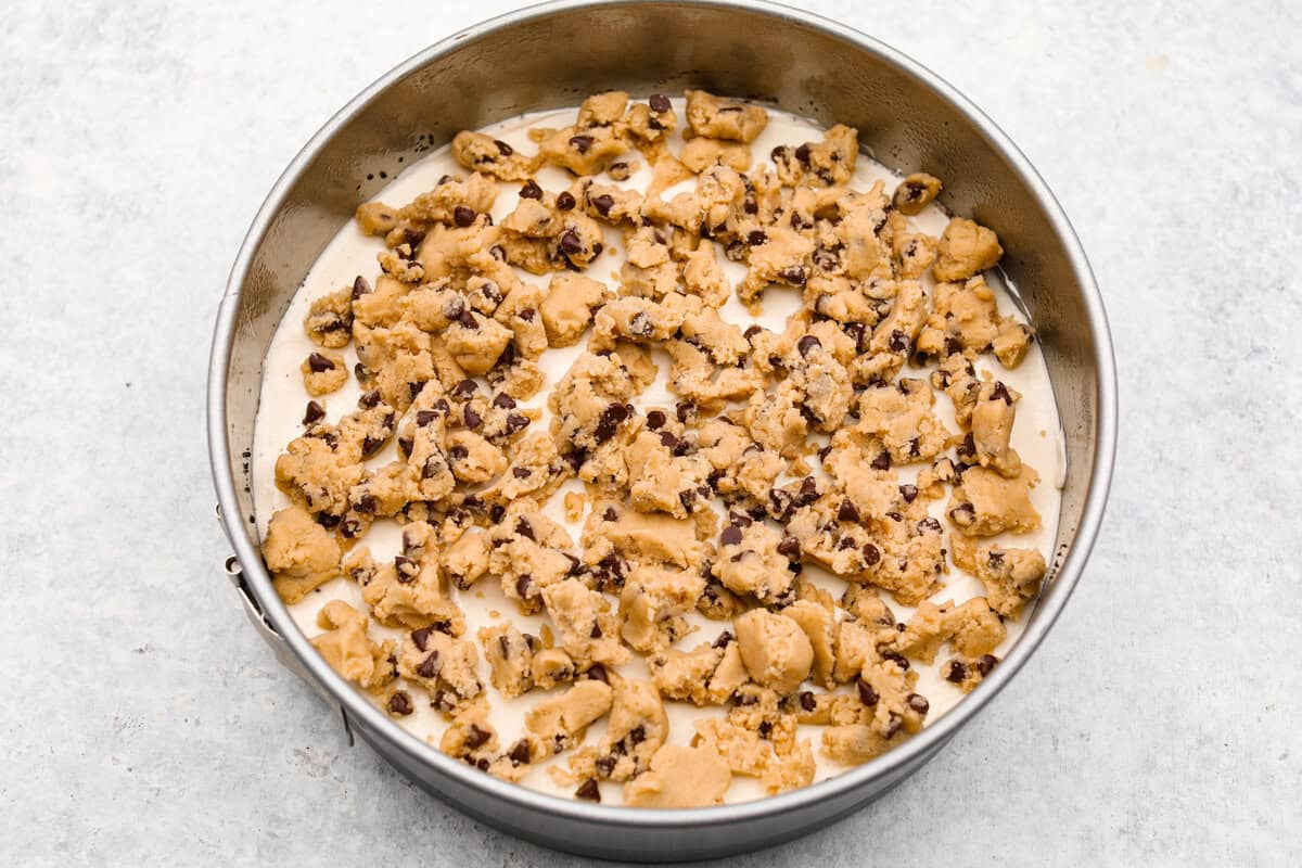 I am so excited about this recipe, it combines two of my all-time favorite desserts, cookie dough and cheesecake! With the rich Oreo crust, creamy cheesecake filling, and edible cookie dough bites, this no-bake cookie dough cheesecake will have your taste buds begging for more! overhead shot of cookie dough pieces on top of the cheesecake layer in a springform pan.