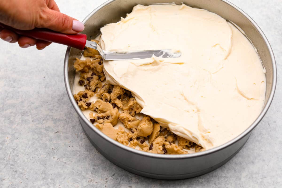 Overhead shot of someone spreading another layer of cheesecake mixture on top of the cookie dough piece layer in the springform pan. 