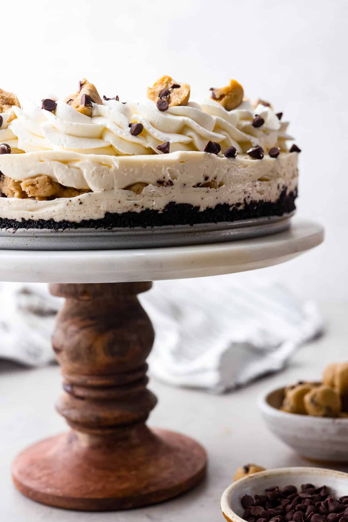 I am so excited about this recipe, it combines two of my all-time favorite desserts, cookie dough and cheesecake! With the rich Oreo crust, creamy cheesecake filling, and edible cookie dough bites, this no-bake cookie dough cheesecake will have your taste buds begging for more! Side shot of completed no-bake cookie dough cheesecake on a cake stand. you can see all the layers and toppings.