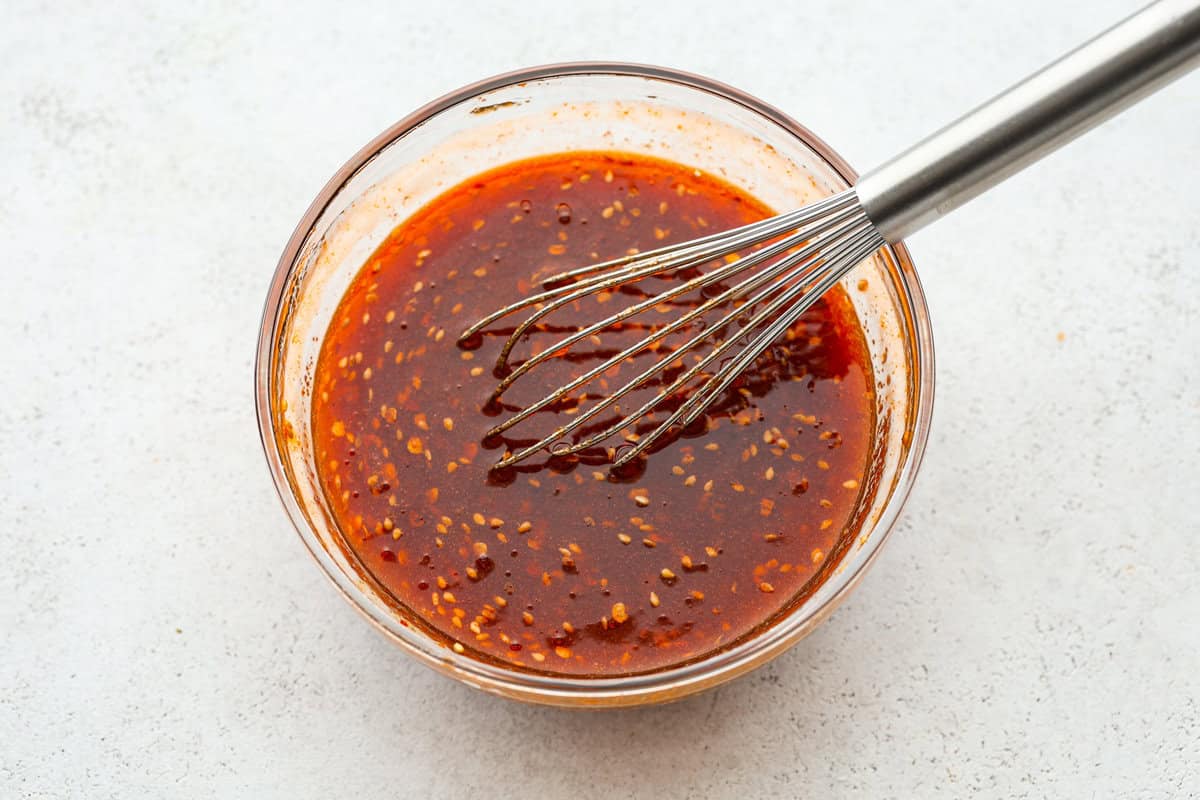 Get ready to spice up your dinner with these firecracker meatballs! Juicy beef meatballs are baked to perfection and then tossed in a sweet and spicy buffalo-based sauce, creating a delicious meal that's perfect for any night of the week. Overhead shot of firecracker sauce with whisk.