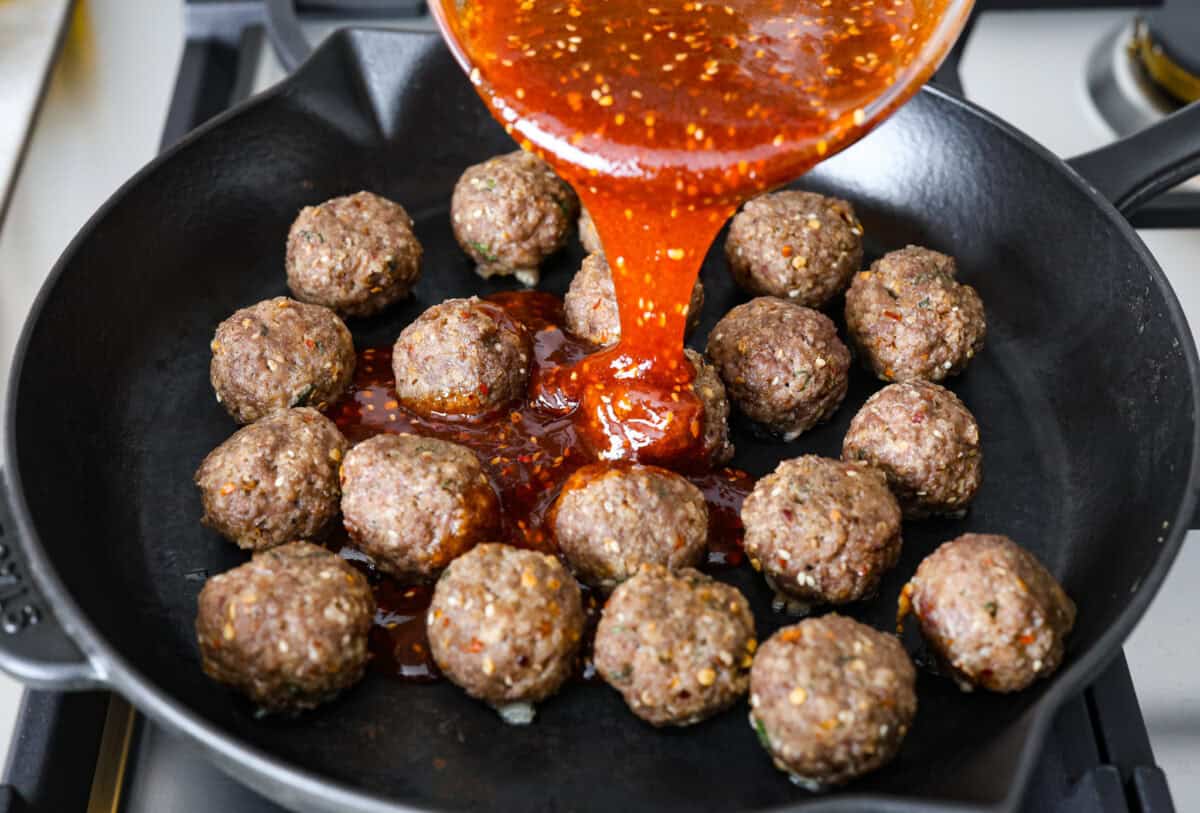 Angle shot of firecracker sauce being poured over cooked meatballs in a skillet. 