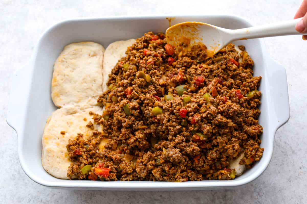This delicious John Wayne Casserole is Mexican comfort food at its finest! Layers of seasoned ground beef, creamy cheddar cheese mixture, and more shredded cheese are baked on top of a biscuit crust for a crowd-pleasing family dinner! The meat mixture being added to the pressed biscuits.
