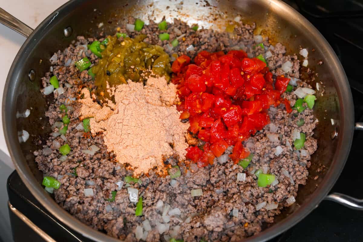 Ground beef in a pan with taco seasoning and tomatoes being added to the meat mixture for John Wayne Casserole. 