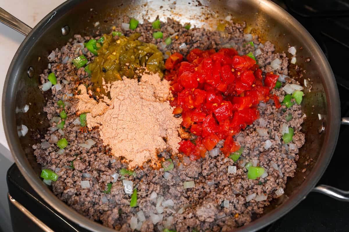 This delicious John Wayne Casserole is Mexican comfort food at its finest! Layers of seasoned ground beef, creamy cheddar cheese mixture, and more shredded cheese are baked on top of a biscuit crust for a crowd-pleasing family dinner! Ground beef in a pan with taco seasoning and tomatoes being added to the meat mixture for John Wayne Casserole.