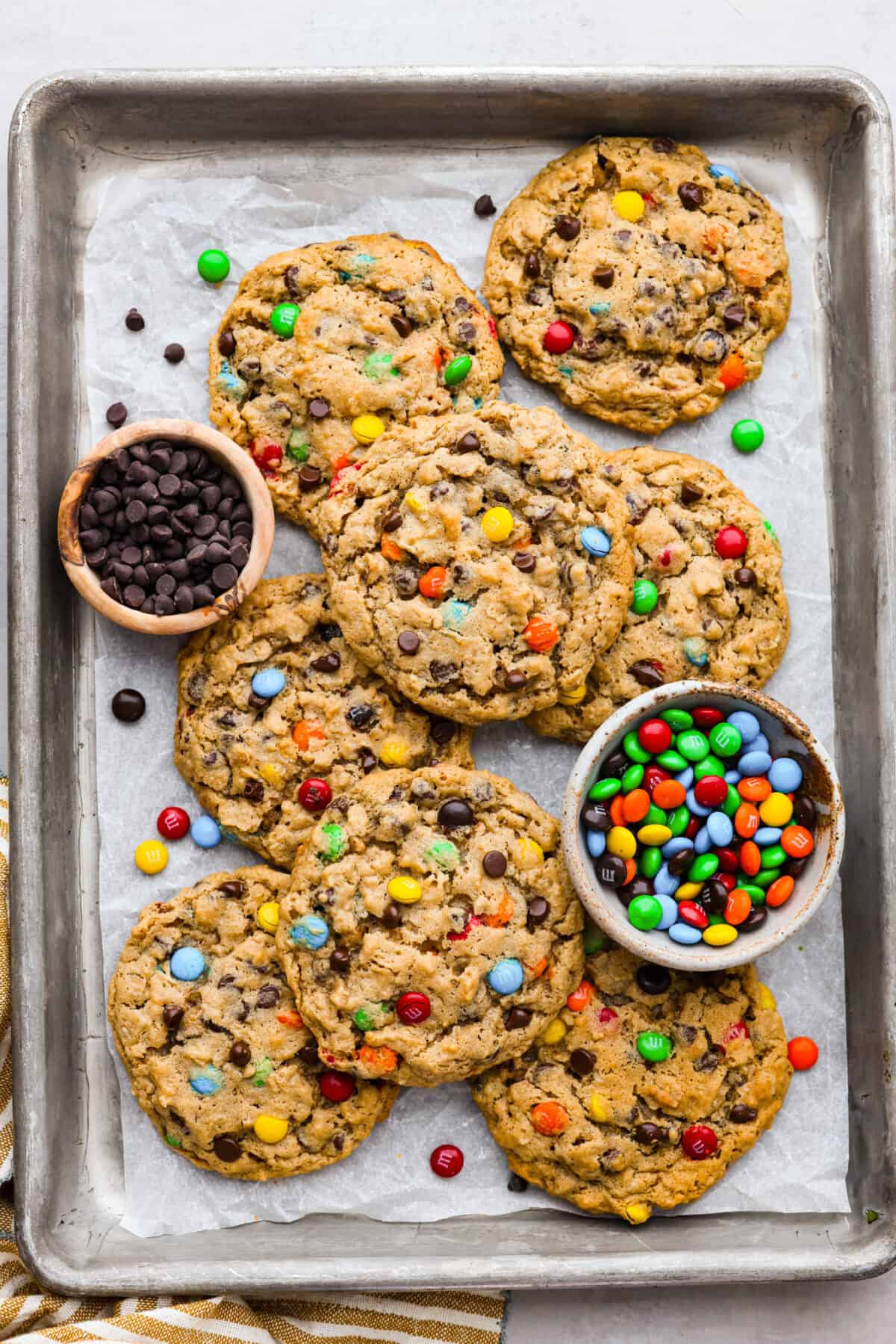 Top view of monster cookies on a sheet pan with bowls of M&M's and chocolate chips.